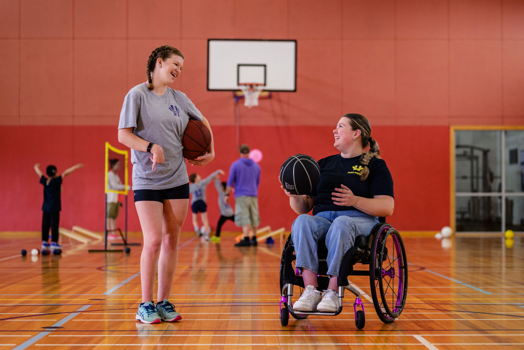 Two women, play basketball in a gym. Women on the right is in a wheelchair
